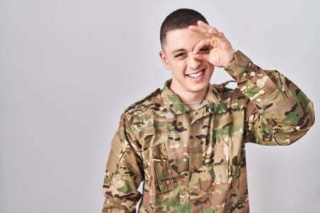 Foto de Joven vestido con camuflaje uniforme del ejército haciendo buen gesto con la mano sonriendo, los ojos mirando a través de los dedos con la cara feliz. - Imagen libre de derechos