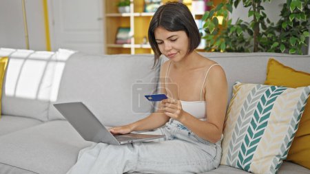 Foto de Joven hermosa mujer hispana de compras con portátil y tarjeta de crédito sentado en el sofá en casa - Imagen libre de derechos