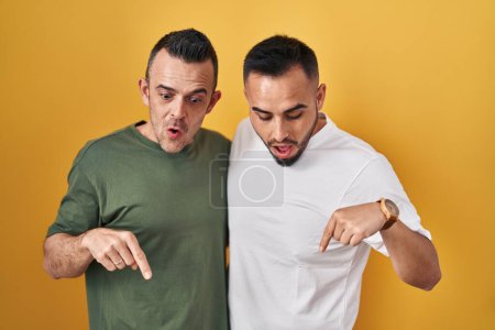Téléchargez les photos : Couple homosexuel debout sur fond jaune pointant vers le bas avec les doigts montrant la publicité, visage surpris et bouche ouverte - en image libre de droit
