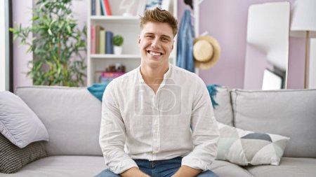 Téléchargez les photos : Joyeux jeune homme caucasien profitant d'un moment de détente, assis avec confiance avec un sourire charmant sur son confortable canapé de salon à la maison - en image libre de droit