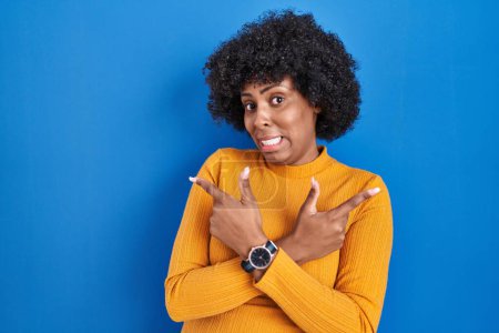 Téléchargez les photos : Femme noire avec les cheveux bouclés debout sur fond bleu pointant vers les deux côtés avec les doigts, direction différente en désaccord - en image libre de droit