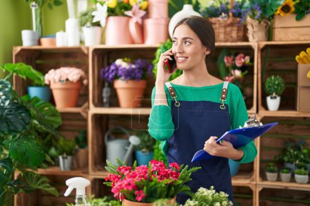 Photo for Young beautiful hispanic woman florist talking on smartphone reading document at flower shop - Royalty Free Image