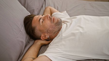 Foto de Retrato cautivador de un apuesto joven hispano agotado, profundamente relajante, dormido, descansando cómodamente, cómodamente acostado en la cama en el interior de su moderno dormitorio - Imagen libre de derechos