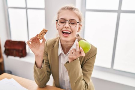 Foto de Joven mujer caucásica trabajando en la oficina comiendo bocadillo sonriendo y riendo en voz alta porque divertida broma loca. - Imagen libre de derechos