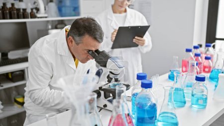 Foto de Científicos masculinos y femeninos trabajando juntos en laboratorio, concentrándose seriamente en su investigación, escribiendo en portapapeles, usando microscopio - Imagen libre de derechos