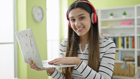 Téléchargez les photos : Jeune belle étudiante hispanique souriante confiante à l'aide d'un ordinateur portable et d'écouteurs à la bibliothèque universitaire - en image libre de droit