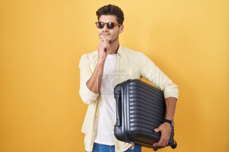 Foto de Joven hombre hispano sosteniendo la maleta yendo de vacaciones de verano mirando confiado a la cámara sonriendo con los brazos cruzados y la mano levantada en la barbilla. pensar positivamente. - Imagen libre de derechos