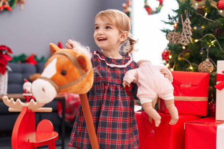 Photo for Adorable caucasian girl holding baby doll and horse toy standing by christmas tree at home - Royalty Free Image