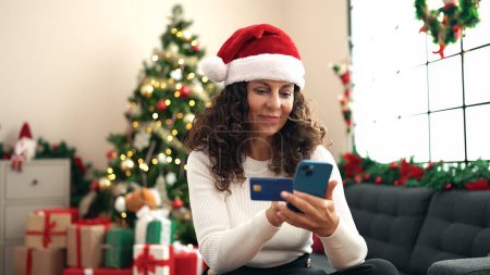 Foto de Mujer hispana de mediana edad usando teléfono inteligente y tarjeta de crédito sentada en el sofá junto al árbol de Navidad en casa - Imagen libre de derechos