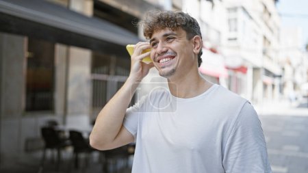 Téléchargez les photos : Jeune homme hispanique écoutant un message vocal souriant sur la terrasse du café - en image libre de droit