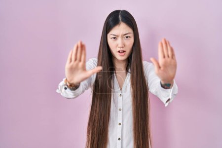 Foto de Mujer joven china de pie sobre fondo rosa haciendo stop gesture con las manos palmas, enojado y expresión de frustración - Imagen libre de derechos