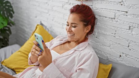 Téléchargez les photos : Jeune belle femme rousse, confortablement détendue en pyjama, souriante tout en utilisant un smartphone sur un lit confortable, profitant d'une matinée calme à l'intérieur dans sa chambre accueillante - en image libre de droit