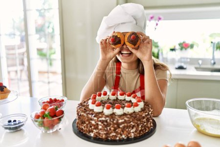 Téléchargez les photos : Jeune belle femme hispanique souriant confiant tenant cupcakes sur les yeux à la cuisine - en image libre de droit