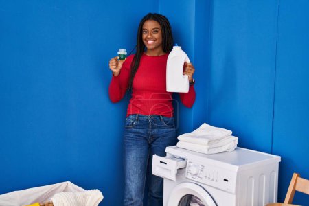 Photo for African american woman smiling confident holding detergent on washing machine at laundry room - Royalty Free Image