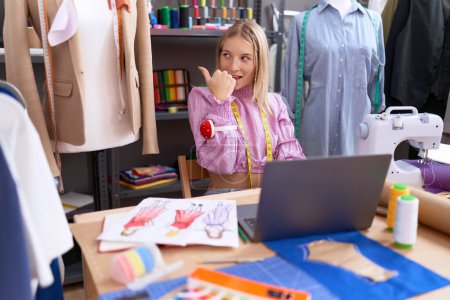 Photo for Young caucasian woman dressmaker designer using laptop pointing thumb up to the side smiling happy with open mouth - Royalty Free Image