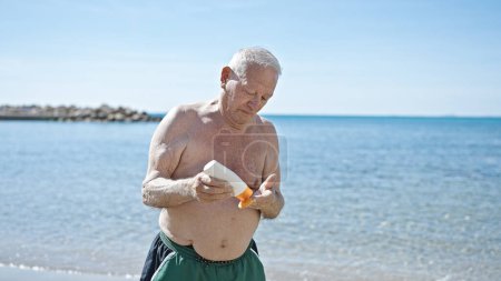 Photo for Senior grey-haired man tourist wearing swimsuit pouring sunscreen on hand at seaside - Royalty Free Image