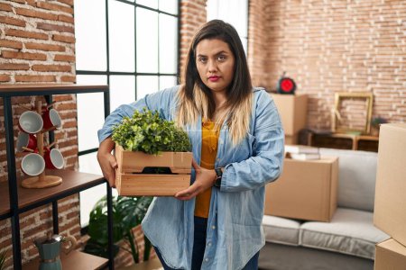Foto de Mujer hispana joven mudándose a un nuevo hogar sosteniendo plantas pensando actitud y expresión sobria buscando confianza en sí misma - Imagen libre de derechos