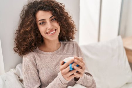 Foto de Mujer hispana joven bebiendo una taza de café sentada en la cama en el dormitorio - Imagen libre de derechos