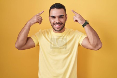 Téléchargez les photos : Jeune homme hispanique debout sur fond jaune souriant pointant vers la tête avec les deux doigts de la main, bonne idée ou pensée, bonne mémoire - en image libre de droit