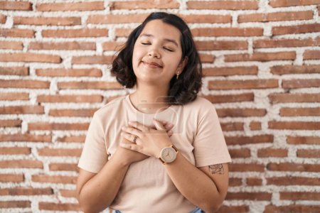 Téléchargez les photos : Jeune femme hispanique debout sur un mur de briques souriant avec les mains sur la poitrine avec les yeux fermés et geste reconnaissant sur le visage. concept de santé. - en image libre de droit