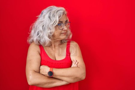 Téléchargez les photos : Femme d'âge moyen avec les cheveux gris debout sur fond rouge regardant sur le côté avec les bras croisés convaincu et confiant - en image libre de droit