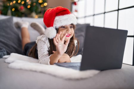 Téléchargez les photos : Adorable hispanique fille ayant appel vidéo couché sur le canapé par arbre de Noël à la maison - en image libre de droit