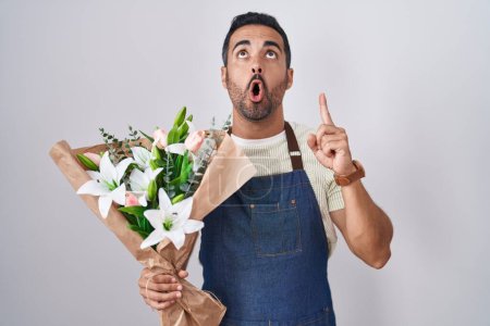 Téléchargez les photos : Homme hispanique avec barbe travaillant comme fleuriste étonné et surpris de regarder vers le haut et pointant avec les doigts et les bras levés. - en image libre de droit