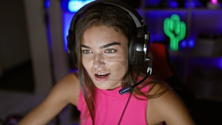 Foto de Atractiva jugadora hispana transmitiendo una sesión de juego nocturno, hablando con sus amigos en línea desde su sala de juegos en casa, disfrutando del entretenimiento virtual mientras se sienta y sonríe en la computadora. - Imagen libre de derechos