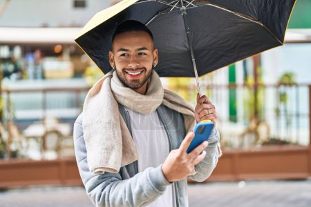 Foto de African american man using smartphone holding umbrella at street - Imagen libre de derechos