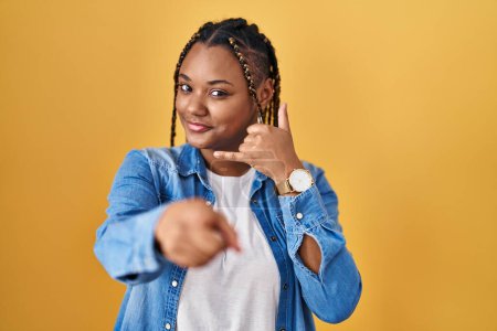 Téléchargez les photos : Femme afro-américaine avec des tresses debout sur fond jaune souriant faire parler au téléphone geste et vous pointant du doigt. Appelle-moi. - en image libre de droit