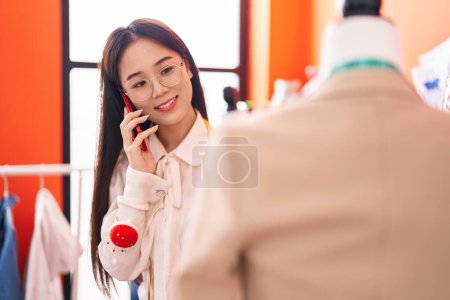 Téléchargez les photos : Jeune femme chinoise tailleur parler sur smartphone regarder veste à l'atelier - en image libre de droit