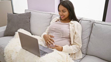 Téléchargez les photos : Radieuse jeune femme enceinte, profitant de son repos maternité, confortablement relaxant à la maison, massant le ventre, souriant positivement à l'écran d'ordinateur portable - en image libre de droit