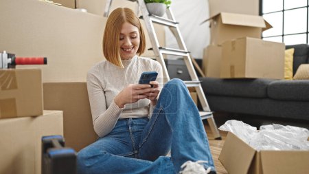 Foto de Mujer rubia joven usando teléfono inteligente sentado en el piso en el nuevo hogar - Imagen libre de derechos