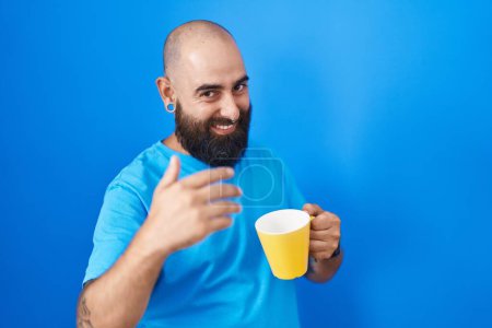 Photo for Young hispanic man with beard and tattoos drinking a cup of coffee beckoning come here gesture with hand inviting welcoming happy and smiling - Royalty Free Image