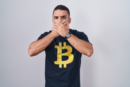 Foto de Joven hombre hispano con camiseta bitcoin impactó cubriendo la boca con las manos por error. concepto secreto. - Imagen libre de derechos
