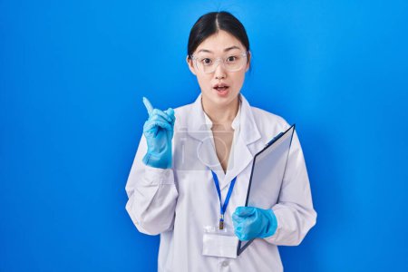 Téléchargez les photos : Chinoise jeune femme travaillant au laboratoire scientifique surpris pointant du doigt sur le côté, bouche ouverte expression étonnée. - en image libre de droit