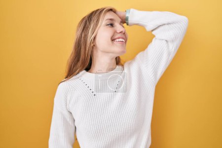 Foto de Mujer joven caucásica vistiendo suéter blanco sobre fondo amarillo sonriendo confiado tocando el pelo con la mano hacia arriba gesto, posando atractivo y de moda - Imagen libre de derechos