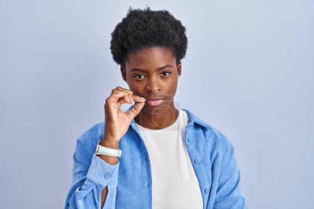 Téléchargez les photos : Femme afro-américaine debout sur fond bleu bouche et lèvres fermées comme zip avec les doigts. secret et silencieux, tabou parlant - en image libre de droit