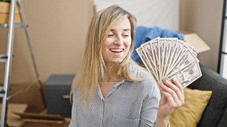 Foto de Joven mujer rubia sonriendo confiado sosteniendo dólares en nuevo hogar - Imagen libre de derechos