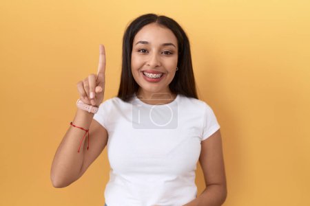 Foto de Mujer árabe joven con camiseta blanca casual sobre fondo amarillo mostrando y apuntando hacia arriba con el dedo número uno mientras sonríe confiado y feliz. - Imagen libre de derechos