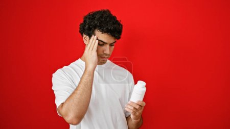 Téléchargez les photos : Jeune homme latin souffrant de maux de tête tenant bouteille de pilules sur fond rouge isolé - en image libre de droit