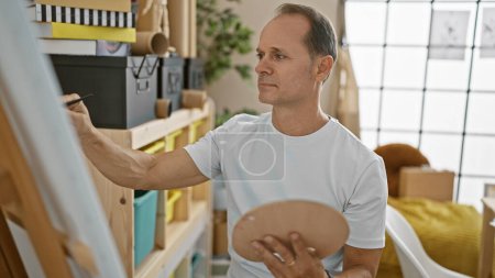 Photo for Confident hispanic middle age man artist drawing enthusiastically on canvas at art studio, surrounded by paintbrushes while sitting relaxed before easel - Royalty Free Image