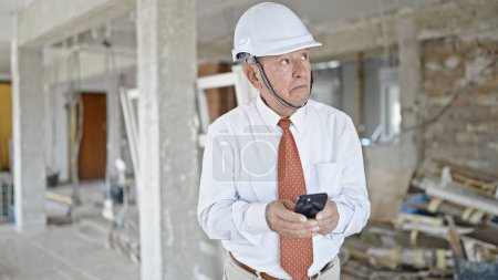 Photo for Senior grey-haired man architect using smartphone looking around at construction site - Royalty Free Image