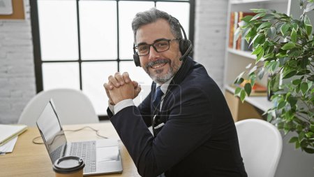 Téléchargez les photos : Homme d'affaires hispanique confiant, jeune et aux cheveux gris, souriant et travaillant au bureau avec son ordinateur portable, à l'écoute du son du succès grâce à son casque. - en image libre de droit