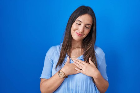 Photo for Young brunette woman standing over blue background smiling with hands on chest with closed eyes and grateful gesture on face. health concept. - Royalty Free Image
