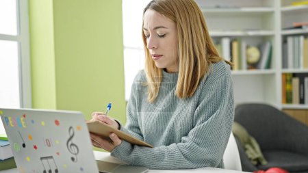 Téléchargez les photos : Jeune étudiante blonde utilisant un ordinateur portable écrivant sur des notes à la bibliothèque de l'université - en image libre de droit