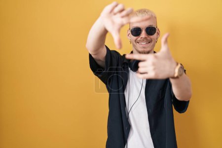 Téléchargez les photos : Jeune homme caucasien portant des lunettes de soleil debout sur fond jaune souriant faisant cadre avec les mains et les doigts avec le visage heureux. concept de créativité et photographie. - en image libre de droit