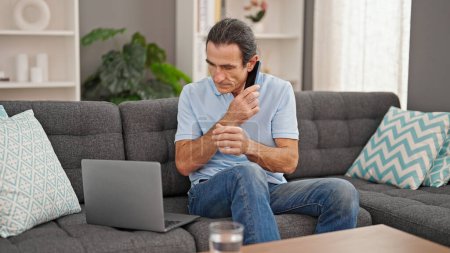 Photo for Middle age man talking on smartphone using laptop at home - Royalty Free Image