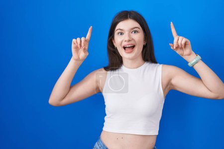 Téléchargez les photos : Jeune femme caucasienne debout sur fond bleu sourire étonné et surpris et pointant vers le haut avec les doigts et les bras levés. - en image libre de droit