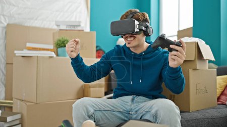 Téléchargez les photos : Jeune homme hispanique jouer à un jeu vidéo en utilisant joystick et lunettes de réalité virtuelle à la nouvelle maison - en image libre de droit